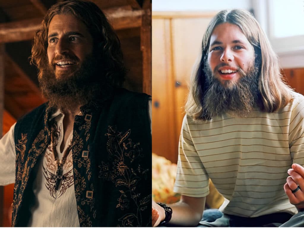 Two men with long hair and bears look straight ahead, one is wearing a t-shirt with brown lines through it, and the other is wearing is a hippie shirt with a beaded cross and a flowered vest. 